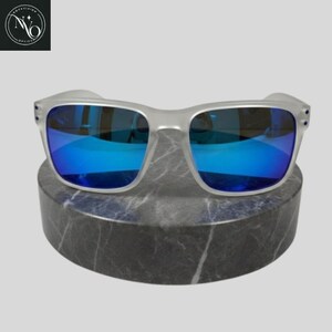 May include: Silver-framed sunglasses with vibrant blue lenses. The sunglasses are displayed on a round, gray marble stand. The lenses reflect a bright blue hue, and the frames have a matte finish. The product is from Novavision Optics.