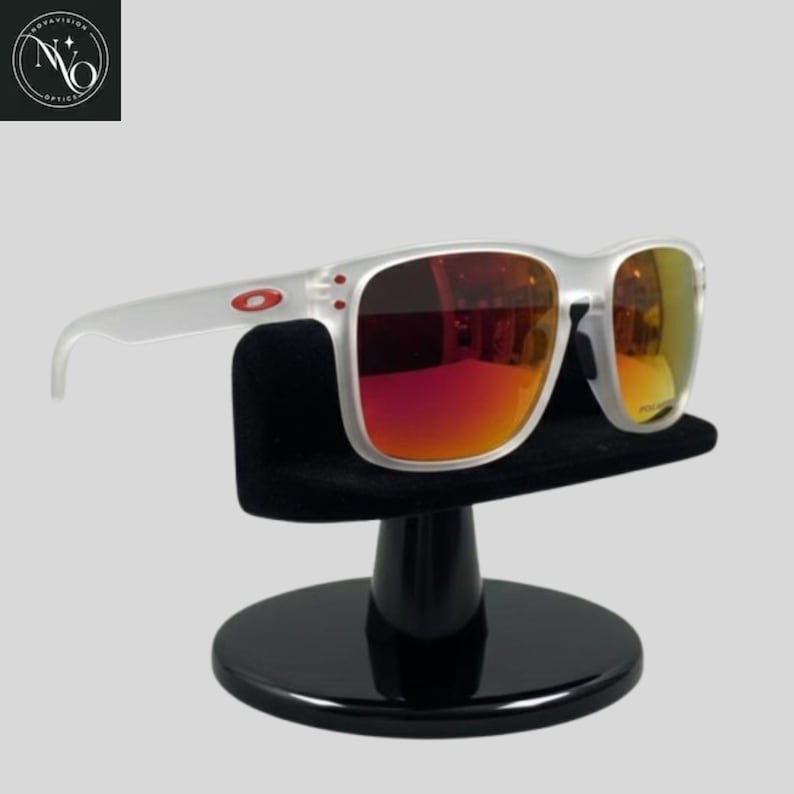 May include: Clear-framed sunglasses with red and yellow mirrored lenses. The sunglasses are displayed on a black stand. The Oakley logo is visible on the temple.