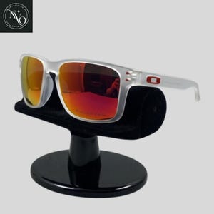 May include: Clear-framed sunglasses with red-orange polarized lenses. The sunglasses are displayed on a black velvet stand. The Oakley logo is visible on the temple.