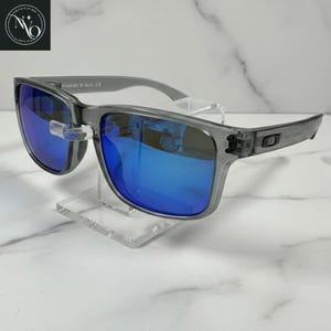 Pu&ograve; includere: Occhiali da sole grigi con lenti blu riflettenti. Gli occhiali hanno una montatura quadrata e sono esposti su un supporto trasparente. Il nome del marchio &egrave; visibile in alto a sinistra.