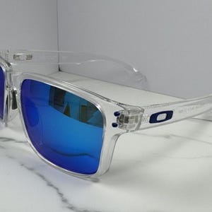 Puede incluir: Gafas de sol con montura transparente y lentes azul brillante. Las gafas tienen forma rectangular y un dise&ntilde;o moderno. Las patillas presentan un peque&ntilde;o logotipo. Las lentes reflejan el entorno, lo que aumenta su atractivo.