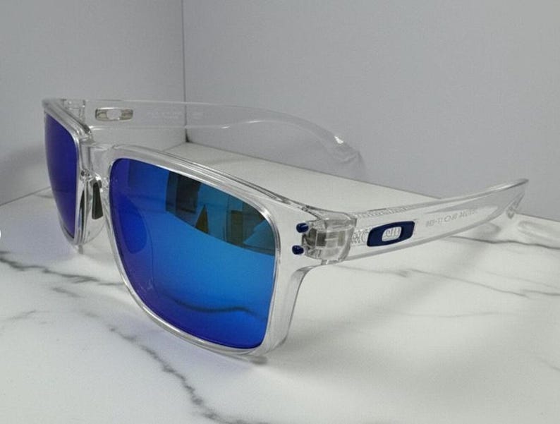 Puede incluir: Gafas de sol con montura transparente y lentes azul brillante. Las gafas tienen forma rectangular y un dise&ntilde;o moderno. Las patillas presentan un peque&ntilde;o logotipo. Las lentes reflejan el entorno, lo que aumenta su atractivo.