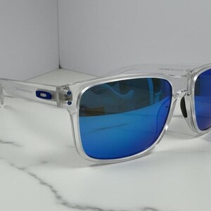 Puede incluir: Gafas de sol con montura transparente y lentes azules espejadas. Las gafas tienen forma cuadrada y un logotipo sutil en la patilla. Las lentes reflejan un color azul brillante y la montura es transparente.