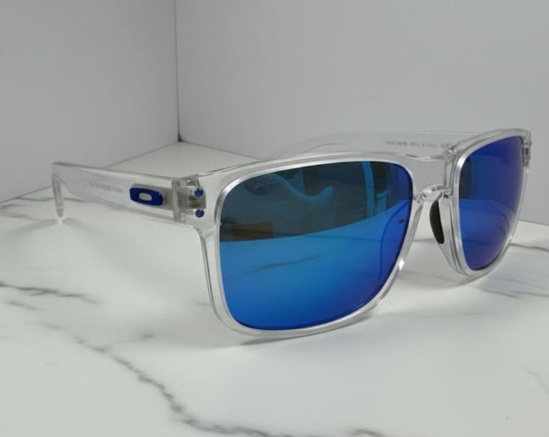Puede incluir: Gafas de sol con montura transparente y lentes azules espejadas. Las gafas tienen forma cuadrada y un logotipo sutil en la patilla. Las lentes reflejan un color azul brillante y la montura es transparente.