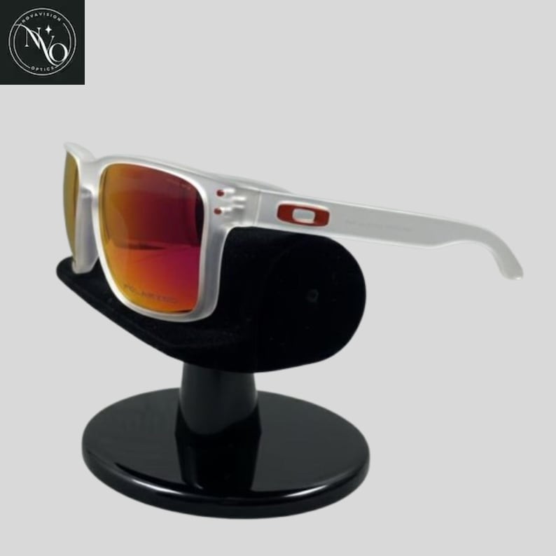May include: White-framed sunglasses with red-tinted lenses are displayed on a black stand. The sunglasses have a rectangular shape and a small red logo on the arm. The stand is round with a black velvet top.