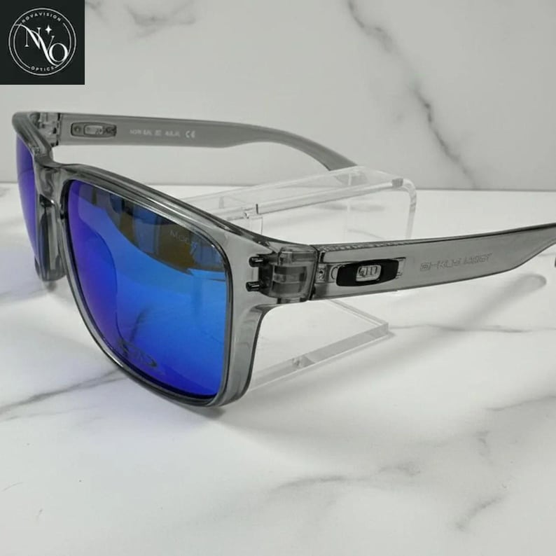 Pu&ograve; includere: Occhiali da sole rettangolari grigi con lenti blu a specchio. Gli occhiali hanno una montatura trasparente e aste grigie. Le lenti riflettono un colore blu brillante. Gli occhiali sono esposti su un supporto trasparente.