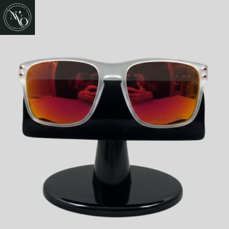 May include: Clear-framed sunglasses with red-tinted lenses are displayed on a black stand. The lenses reflect the surroundings. The sunglasses have a rectangular shape and are labeled "POLARIZED". The stand has a round base.