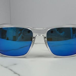Puede incluir: Gafas de sol con montura transparente y lentes espejadas azules vibrantes. Las monturas cuadradas tienen un dise&ntilde;o moderno, que refleja el entorno. Las gafas de sol est&aacute;n sobre una superficie de m&aacute;rmol blanco.