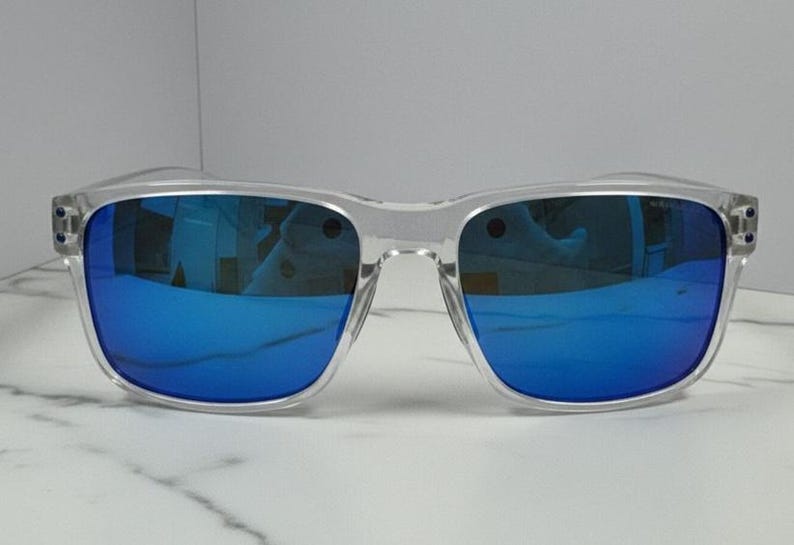 Puede incluir: Gafas de sol con montura transparente y lentes espejadas azules vibrantes. Las monturas cuadradas tienen un dise&ntilde;o moderno, que refleja el entorno. Las gafas de sol est&aacute;n sobre una superficie de m&aacute;rmol blanco.
