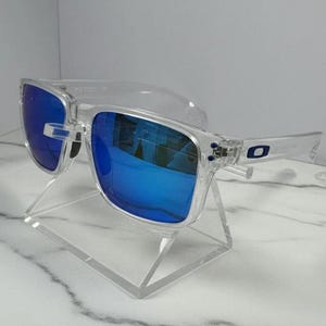 Puede incluir: Gafas de sol con montura transparente y lentes azul brillante. Las gafas de sol rectangulares se presentan sobre un soporte de acr&iacute;lico transparente. Las lentes reflejan el entorno, creando un efecto visual din&aacute;mico.