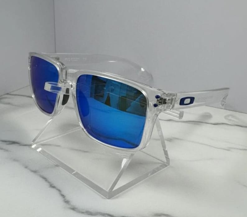 Puede incluir: Gafas de sol con montura transparente y lentes azul brillante. Las gafas de sol rectangulares se presentan sobre un soporte de acr&iacute;lico transparente. Las lentes reflejan el entorno, creando un efecto visual din&aacute;mico.