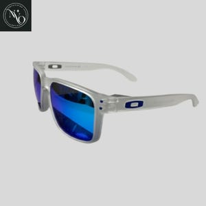 May include: Clear-framed sunglasses with blue mirrored lenses. The sunglasses have a rectangular shape and white arms. The Oakley logo is visible on the temples. The lenses reflect a bright blue color.