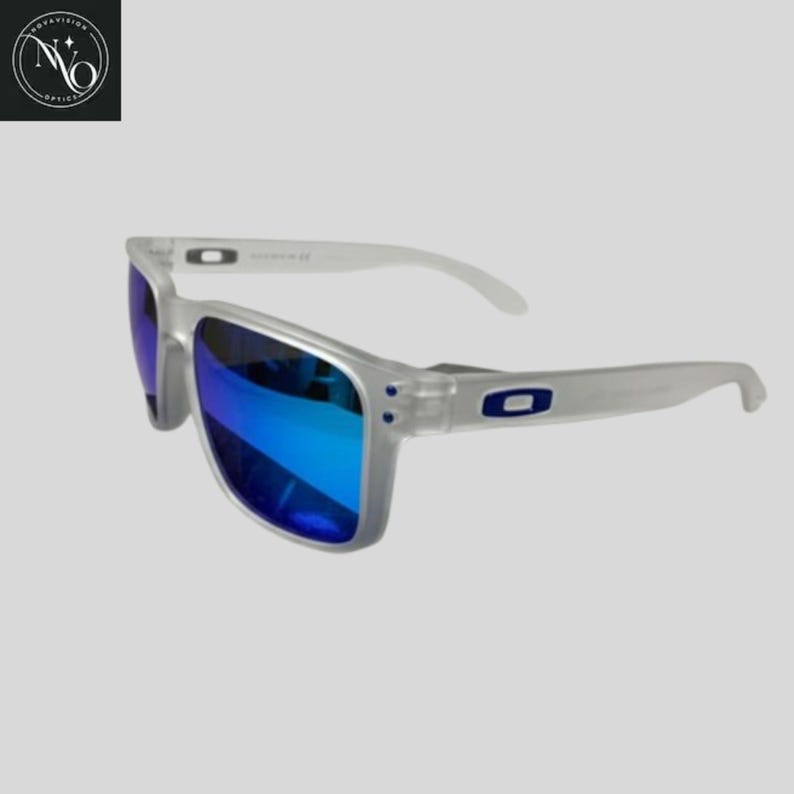 May include: Clear-framed sunglasses with blue mirrored lenses. The sunglasses have a rectangular shape and white arms. The Oakley logo is visible on the temples. The lenses reflect a bright blue color.