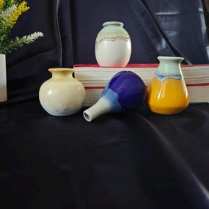 Puede incluir: Una colección de jarrones de cerámica en varios colores y formas. Los jarrones incluyen un jarrón blanco con un borde marrón y rojo, un jarrón amarillo, un jarrón azul y un jarrón amarillo y azul. Los jarrones están dispuestos sobre una superficie oscura.