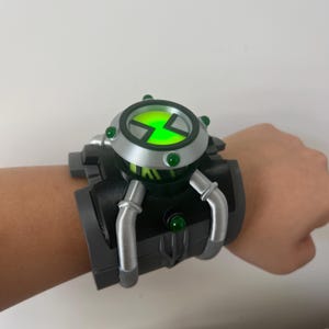 Op de afbeelding: Een grijs en zwart polsapparaat met een cirkelvormige bovenkant, een oplichtend neongroen midden en een zwart zandlopervormig symbool. Het accessoire bevat zilverkleurige accenten, kleine groene ronde noppen en gebogen zilveren buizen.