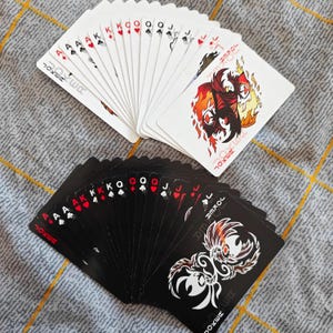 May include: Two decks of playing cards, one black and one white, fanned out on a patterned surface. The white deck shows red and black suits, while the black deck has white suits. Each deck has a Joker card with a unique design.