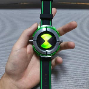 May include: A black and green wristwatch featuring a circular face with a silver metallic bezel and a central glowing green hourglass symbol. The watch band is black with a thin green stripe running down the center of the strap.