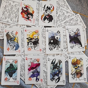 Puede incluir: Una baraja de cartas personalizada con ilustraciones únicas. Las cartas muestran diseños de personajes coloridos, con las cartas Joker que muestran figuras de dos tonos. Los dorsos de las cartas tienen un patrón floral decorativo.