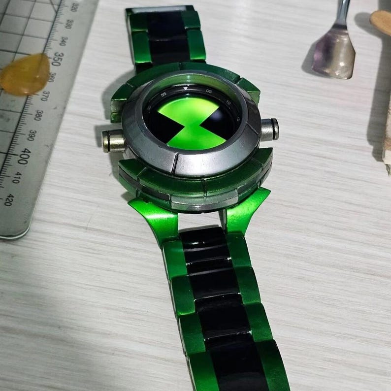 May include: A metallic green and black wristwatch featuring a circular face with a glowing green hourglass symbol in the center. The watch band consists of alternating green and black rectangular links, and a metal ruler is visible to the left.