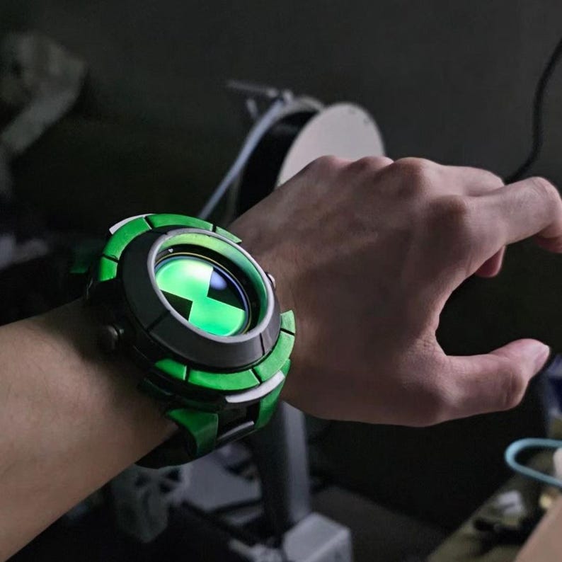 May include: A chunky wrist-worn device featuring a circular face with a glowing green hourglass symbol. The casing is primarily black with bright green accents along the outer rim, designed with a segmented, mechanical aesthetic and a thick strap.