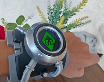 May include: A wrist-worn device featuring a black and silver frame with a circular center. The center displays a bright green diamond-shaped icon with a black graphic symbol inside. The device has a thick, structured band with silver accents.