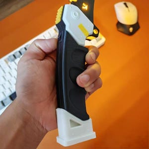 Op de afbeelding: Een zwart-wit handheld apparaat met gele accenten wordt vastgehouden. Het apparaat heeft een getextureerde grip en een witte basis. Een toetsenbord en muis zijn zichtbaar op de achtergrond.
