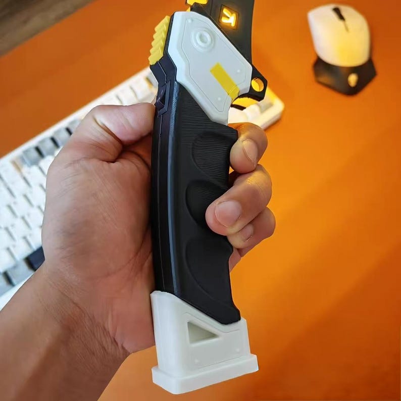 Op de afbeelding: Een zwart-wit handheld apparaat met gele accenten wordt vastgehouden. Het apparaat heeft een getextureerde grip en een witte basis. Een toetsenbord en muis zijn zichtbaar op de achtergrond.