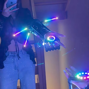 Puede incluir: Un accesorio futurista para brazo y mano con luces LED integradas. Las luces están dispuestas en filas y círculos, emitiendo una vibrante gama de colores. El accesorio es negro y plateado, con un diseño elegante y tecnológico.