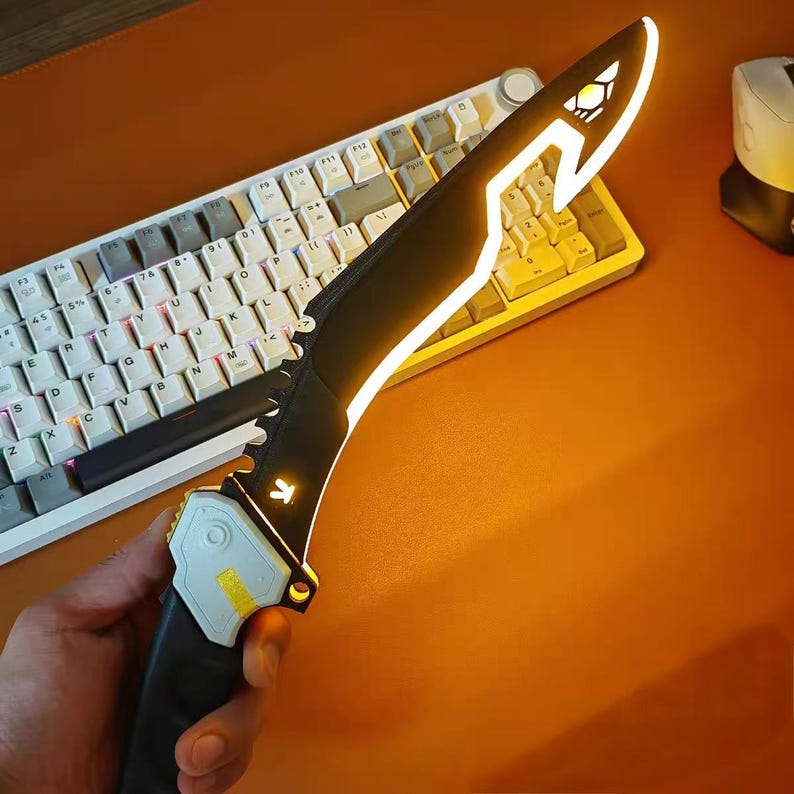 Op de afbeelding: Een zwart en geel LED-mes met een uniek ontwerp. Het lemmet heeft een gloeiende gele omtrek en een gestileerde vorm. De handgreep heeft een wit en geel accent. Het mes ligt op een toetsenbord.