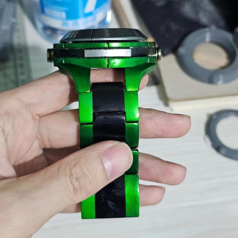 May include: A wristwatch featuring a metallic green finish on the case and outer bracelet links, contrasted by a central row of black links. The watch has a silver-toned accent band around the side of the case and a dark, flat watch face.