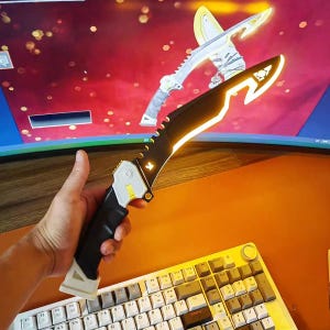 Puede incluir: Un cuchillo plegable negro y amarillo con borde dentado y hoja curva se sostiene en una mano. El cuchillo tiene un mango negro y un contorno amarillo brillante. Un segundo cuchillo es visible en el fondo.