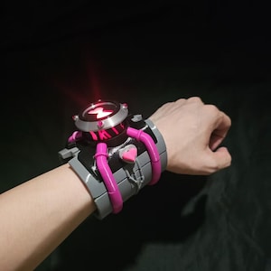 May include: A wrist-worn device featuring a black and grey cuff with bright pink accents and a central circular dial. The dial displays a glowing red light with a heart-shaped symbol. The structure includes pink tubing details and a small heart icon.