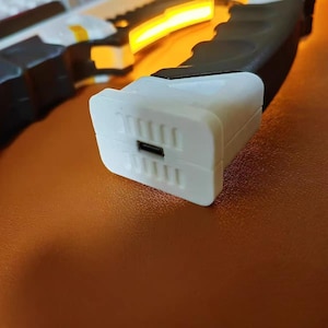 Op de afbeelding: Een close-up van een wit rechthoekig object met een zwarte USB-poort en verticale groeven. Het object maakt deel uit van een groter zwart-wit apparaat met gele accenten en een helder geel licht.