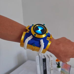 Puede incluir: Un dispositivo de muñeca con un dial circular central que emite una luz azul brillante, rodeado por un marco dorado y azul. El accesorio incluye detalles curvos en blanco y pequeños elementos circulares en color verde azulado.