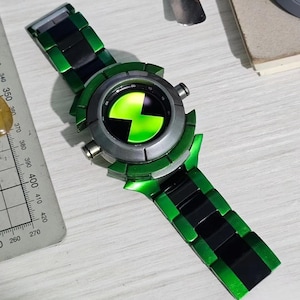 May include: A wristwatch featuring a metallic green and black link band and a circular face with a bright green and black hourglass-shaped symbol. The watch is positioned next to a metal ruler showing measurements in millimetres.