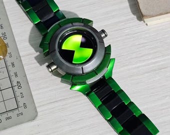 May include: A wristwatch featuring a metallic green and black link band and a circular face with a bright green and black hourglass-shaped symbol. The watch is positioned next to a metal ruler showing measurements in millimeters.