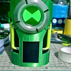 May include: A green and silver wrist-worn device featuring a circular dial with a central hourglass symbol. The structure includes bright yellow accents on the sides and a black rectangular panel on the front, positioned over a measuring mat.