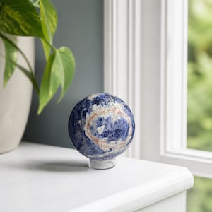 May include: A polished spherical stone ornament featuring a deep blue base color with prominent white and reddish-brown mineral inclusions. The sphere rests securely on a small, clear, circular acrylic stand, showcasing its natural stone texture.