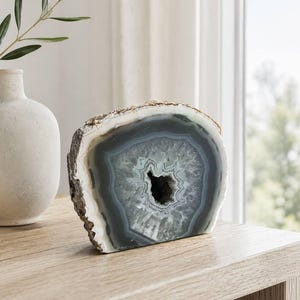May include: A polished agate slice featuring concentric bands of gray, white, and translucent blue tones surrounding a central crystalline geode cavity. The outer edge of the stone retains a natural, rough, and textured brown mineral crust.