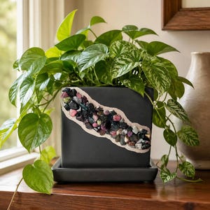 May include: A square matte black ceramic planter with an attached saucer, featuring a diagonal decorative inlay of multi-colored raw tourmaline crystals in shades of pink, green, and black, containing a trailing green pothos plant with variegated leaves.
