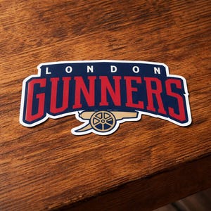 May include: A sticker with the words "LONDON GUNNERS" in red, outlined in white, on a navy blue background. A gold cannon graphic is below the text. The sticker is on a wooden surface.