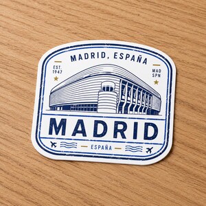 Puede incluir: Una pegatina cuadrada con esquinas redondeadas que presenta una ilustraci&oacute;n de l&iacute;neas azules de un estadio. El texto dice MADRID, ESPA&Ntilde;A, EST. 1947, MAD SPN y MADRID. El dise&ntilde;o incluye peque&ntilde;as estrellas doradas, iconos de aviones y l&iacute;neas onduladas.