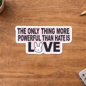 May include: A rectangular sticker with the text "THE ONLY THING MORE POWERFUL THAN HATE IS LOVE" in black letters on a pink background. The word "LOVE" features a bunny face. The sticker is on a wooden surface with a small plant, a pencil, and a notepad.