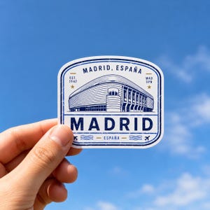 Puede incluir: Una pegatina blanca con una ilustraci&oacute;n gr&aacute;fica azul de un estadio en Madrid, Espa&ntilde;a. El texto dice MADRID, ESPA&Ntilde;A, EST. 1947, MAD SPN y MADRID. El dise&ntilde;o presenta un borde de estilo vintage desgastado con peque&ntilde;as estrellas e iconos de aviones.
