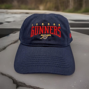 May include: Navy blue baseball cap with the text "LONDON GUNNERS" embroidered in red, with a gold cannon emblem. The cap has a curved brim and a structured crown. The cap is made of a durable fabric.