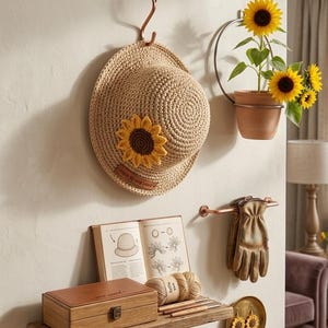 Puede incluir: Un sombrero de paja con un girasol de ganchillo cuelga de una pared. Una pequeña maceta de terracota con girasoles está montada cerca. Una estantería de madera contiene una caja de madera, un libro, lana y ganchos de ganchillo.