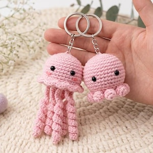 May include: Two light pink crochet keychains featuring a jellyfish and an octopus. Both items have black bead eyes and are attached to silver metal key rings with short chains. The jellyfish has long, spiraled tentacles, while the octopus has short, curled arms.