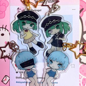 Puede incluir: Cuatro llaveros acrílicos transparentes con personajes de estilo anime. Dos tienen cabello verde y visten trajes de marinero, mientras que los otros dos tienen cabello azul y trajes de baño. Un personaje de cabello azul tiene el texto "Do you TRUST ME?" Llaveros dorados en forma de estrella.