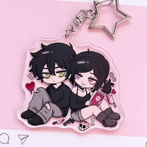 May include: An acrylic keyring featuring a cartoon illustration of two characters with black hair sitting together. The design includes a red heart, a skull, a book with a skull icon, a knife, and a star-shaped metal clasp attached to the top.