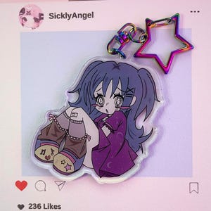 May include: A clear acrylic charm depicting an anime-style girl with blue hair and a purple dress. The charm has a rainbow-coloured star-shaped keyring and a matching clasp. The image is on a pink background with an Instagram interface.